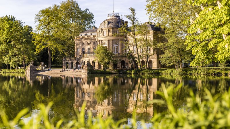 Schloss Monrepos spiegelt sich im ruhigen Wasser eines Sees, umgeben von &uuml;ppigem Gr&uuml;n und B&auml;umen. Menschen spazieren am Ufer entlang., &copy; SMG, Werner Dieterich