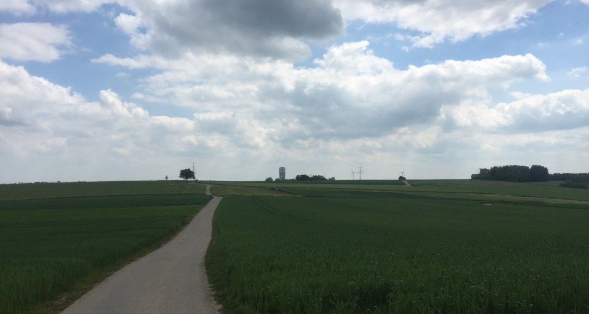 Ein schmaler Weg führt durch grüne Felder zu einem fernen Wasserturm und Windrädern, unter einem bewölkten Himmel., © www.pro-cycl.de Ein schmaler Weg führt durch grüne Felder zu einem fernen Wasserturm und Windrädern, unter einem bewölkten Himmel., © www.pro-cycl.de