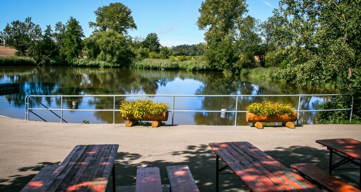 Ein idyllischer See mit Bäumen im Hintergrund, umgeben von Natur. Im Vordergrund stehen Bänke und Blumenkästen mit gelben Blumen., © agentur arcos/Niki Eilers Ein idyllischer See mit Bäumen im Hintergrund, umgeben von Natur. Im Vordergrund stehen Bänke und Blumenkästen mit gelben Blumen., © agentur arcos/Niki Eilers