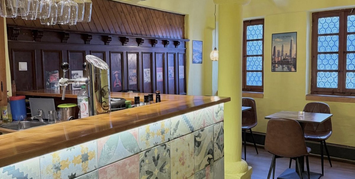 Ein gem&uuml;tliches Restaurant mit gelben W&auml;nden, einer Bar mit bunten Fliesen und h&auml;ngenden Weingl&auml;sern. Im Hintergrund sind Tische und St&uuml;hle zu sehen.