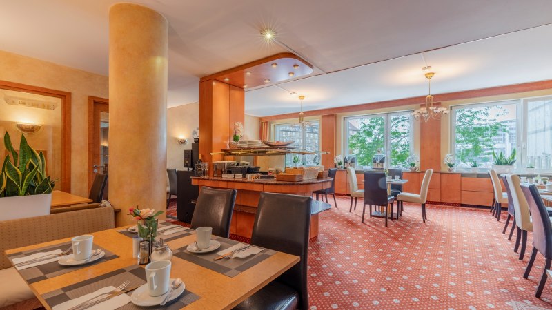 Eleganter Fr&uuml;hst&uuml;cksraum mit gedeckten Tischen, Pflanzen und gro&szlig;en Fenstern. Warme Farben und gem&uuml;tliche Atmosph&auml;re., &copy; PLAZA INN Rieker Stuttgart Hauptbahnhof