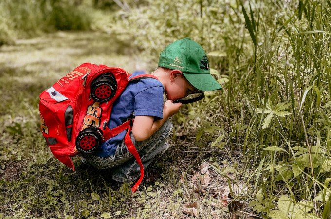 Ein Kind mit rotem Rucksack und gr&uuml;ner Kappe erkundet mit einer Lupe die Pflanzenwelt auf einer Wiese., &copy; Stadt N&uuml;rtingen