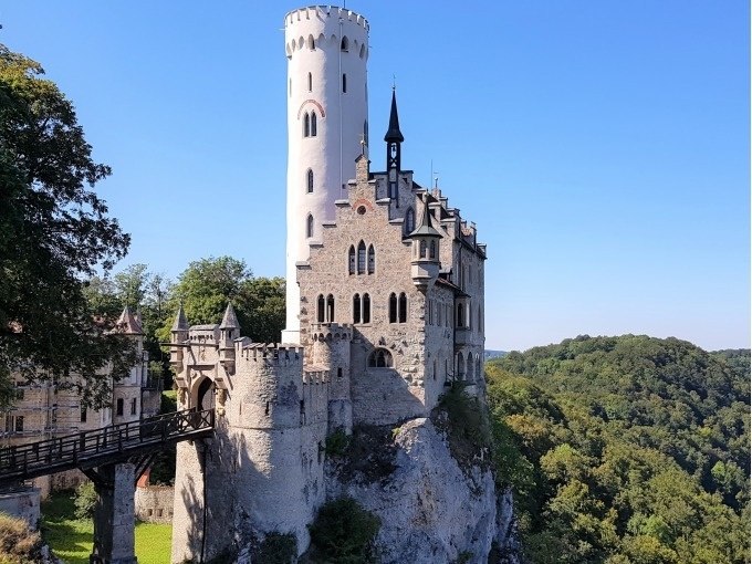 Schloss Lichtenstein thront majest&auml;tisch auf einem Felsen, umgeben von Wald und unter klarem, blauem Himmel., &copy; SwabianTravel