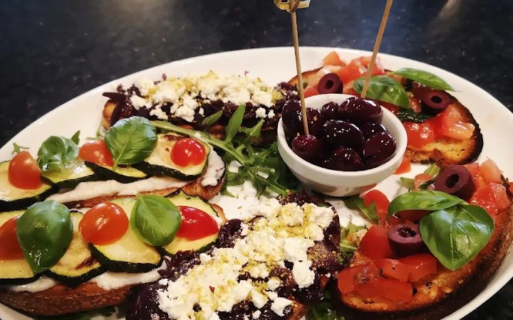 Ein Teller mit Antipasti: Bruschetta mit Tomaten, Zucchini, Oliven und Basilikum, dazu eine Schale mit Oliven in der Mitte., © La buona Forketta