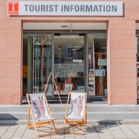 Die TOURIST INFORMATION mit Glasfront und zwei Liegest&uuml;hlen vor dem Eingang. Vor dem Geb&auml;ude stehen Blumen und ein Holzstamm., &copy; Touristik Bad Wildbad GmbH
