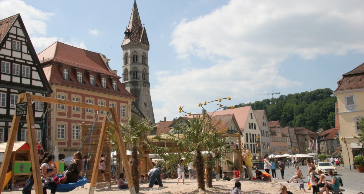 Sandspielplatz mit Nestschaukel, Palmen und vielen Menschen. Im Hintergrund Häuser der Stadt und der Johannisturm., © Foto: Cornelia Steinbach Sandspielplatz mit Nestschaukel, Palmen und vielen Menschen. Im Hintergrund Häuser der Stadt und der Johannisturm., © Foto: Cornelia Steinbach
