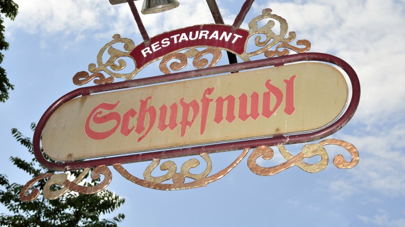 Ein dekoratives Schild mit der Aufschrift 'Schupfnudl' hängt vor einem blauen Himmel. Es gehört zu einem Restaurant., © Schupfnudl Ein dekoratives Schild mit der Aufschrift 'Schupfnudl' hängt vor einem blauen Himmel. Es gehört zu einem Restaurant., © Schupfnudl