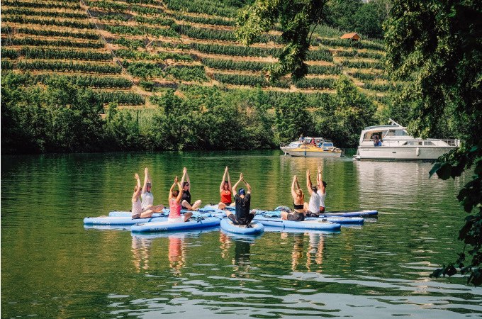 Menschen praktizieren Yoga auf SUP-Boards auf einem Fluss, umgeben von Weinbergen und einem vorbeifahrenden Boot., &copy; Cool-Tours StattReisen
