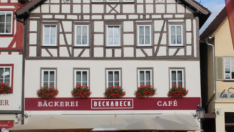 Fachwerkhaus mit Bäckerei und Café Beckabeck. Rote Blumen schmücken die Fenster. Vor dem Gebäude stehen Tische und Stühle unter Sonnenschirmen.