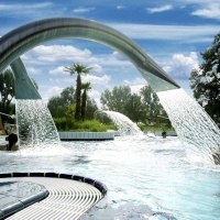 Ein Außenpool mit Wasserfontänen und Palmen, umgeben von Bäumen und einem Gebäude. Menschen genießen das Wasser unter blauem Himmel., © Stuttgarter Bäder