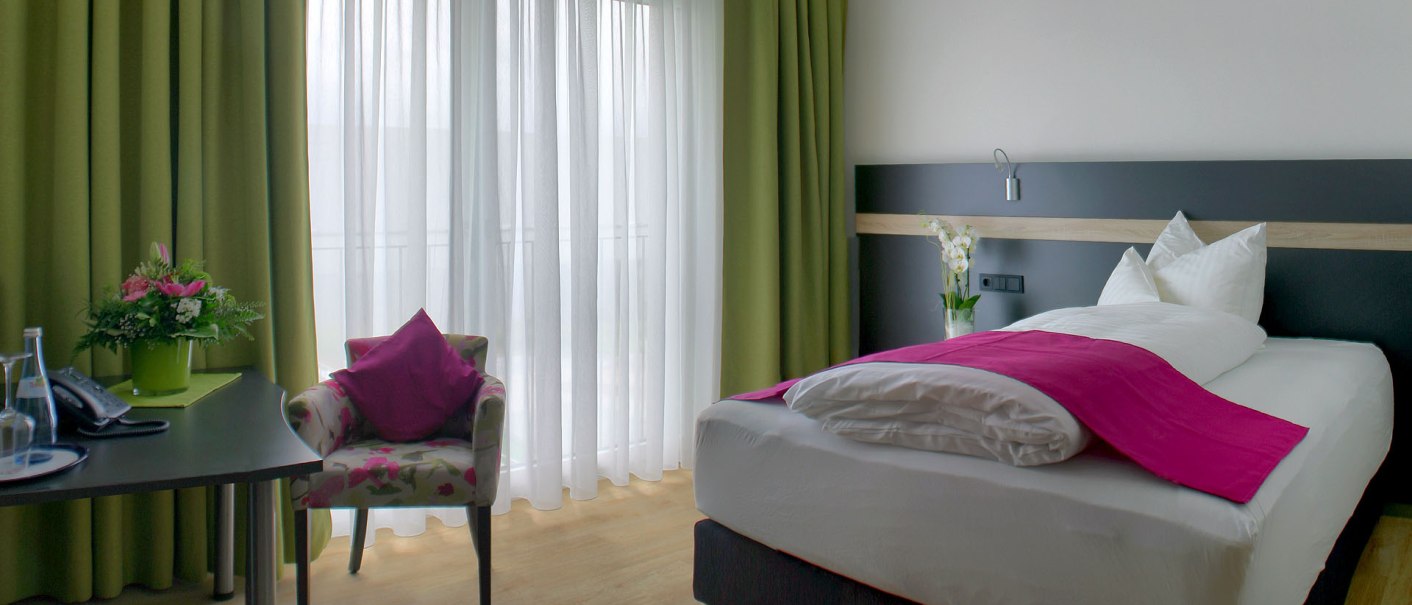 Ein modernes Einzelzimmer mit einem Bett, einem Tisch mit Blumen, einem Stuhl und grünen Vorhängen vor einem Fenster., © TOMAS