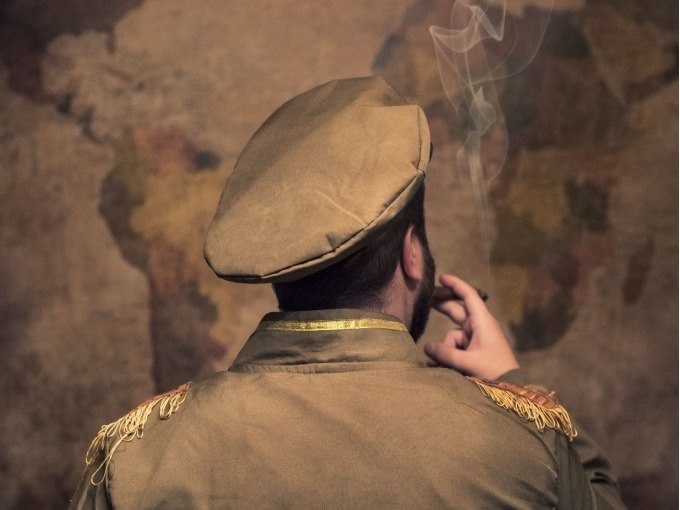 Person in Militäruniform mit Mütze raucht vor einer Weltkarte. Der Hintergrund ist unscharf, der Rauch ist sichtbar., © ExitGames Stuttgart Person in Militäruniform mit Mütze raucht vor einer Weltkarte. Der Hintergrund ist unscharf, der Rauch ist sichtbar., © ExitGames Stuttgart