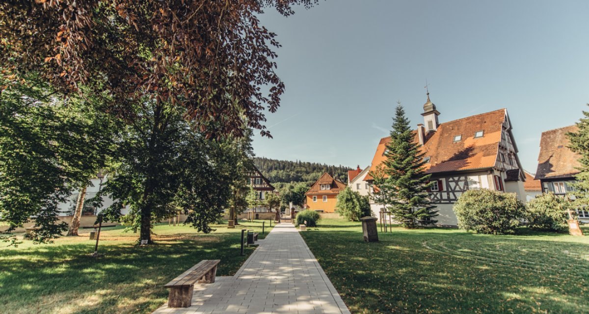 Gepflasterter Weg durch grünen Garten mit Bäumen und Bänken, umgeben von Fachwerkhäusern und Kirchturm im Hintergrund., © Tourismus Ostalb Gepflasterter Weg durch grünen Garten mit Bäumen und Bänken, umgeben von Fachwerkhäusern und Kirchturm im Hintergrund., © Tourismus Ostalb
