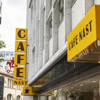 Gelbes Schild des Cafe Nast mit Kaffeetasse-Logo an einem Gebäude, umgeben von Bäumen., © SMG, Sarah Schmid