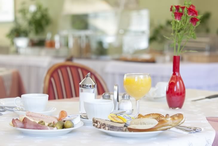 Fr&uuml;hst&uuml;ckstisch mit Br&ouml;tchen, Aufschnitt, Orangensaft und einer roten Vase mit Blumen in einem hellen Raum., &copy; Hotel Drei Morgen