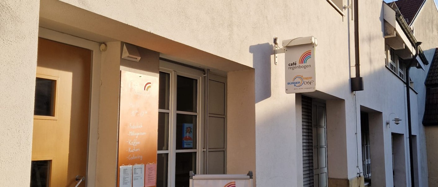 Das Bild zeigt die Au&szlig;enansicht des Caf&eacute; Regenbogen mit einem Schild und einem Aufsteller vor der Eingangst&uuml;r. Die Fassade ist hell und die Stra&szlig;e gepflastert., &copy; cStadtNuertingen