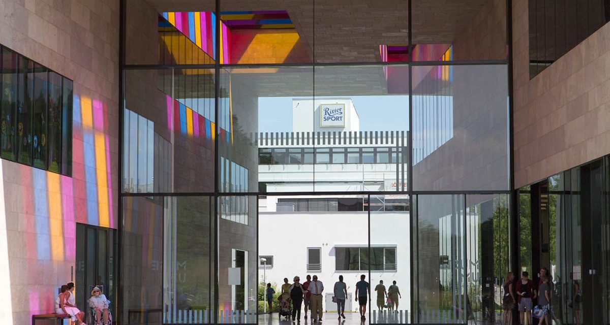 Eingangsbereich des Museum Ritter mit bunten Lichtreflexionen auf den Wänden. Menschen gehen durch den Glasdurchgang, im Hintergrund das Ritter Sport Logo., © Stuttgart Marketing GmbH Eingangsbereich des Museum Ritter mit bunten Lichtreflexionen auf den Wänden. Menschen gehen durch den Glasdurchgang, im Hintergrund das Ritter Sport Logo., © Stuttgart Marketing GmbH