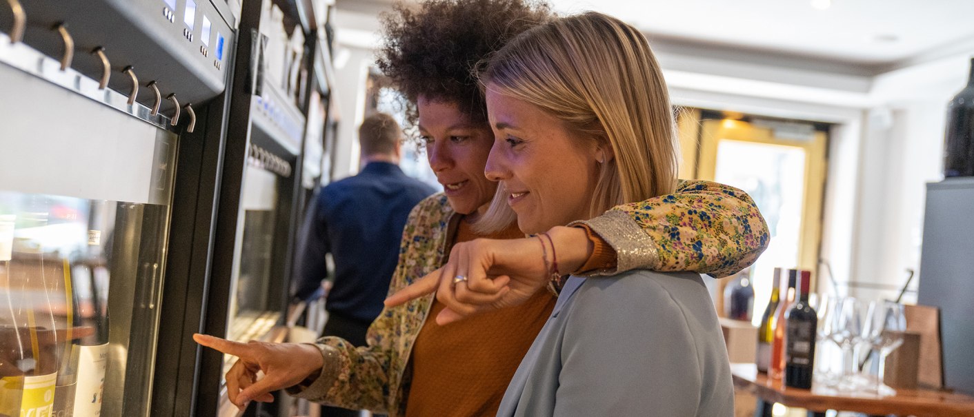 Zwei Frauen stehen vor einem Weinspender und zeigen auf verschiedene Weinflaschen. Im Hintergrund sind weitere Flaschen und Gläser zu sehen., © Stuttgart Marketing GmbH, Fotografin Martina Denker Zwei Frauen stehen vor einem Weinspender und zeigen auf verschiedene Weinflaschen. Im Hintergrund sind weitere Flaschen und Gläser zu sehen., © Stuttgart Marketing GmbH, Fotografin Martina Denker