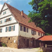 Fachwerkhaus mit rotem Dach und blauen Fensterläden, umgeben von Bäumen. Das Gebäude steht auf einem steinernen Fundament., © Alfred Rösner