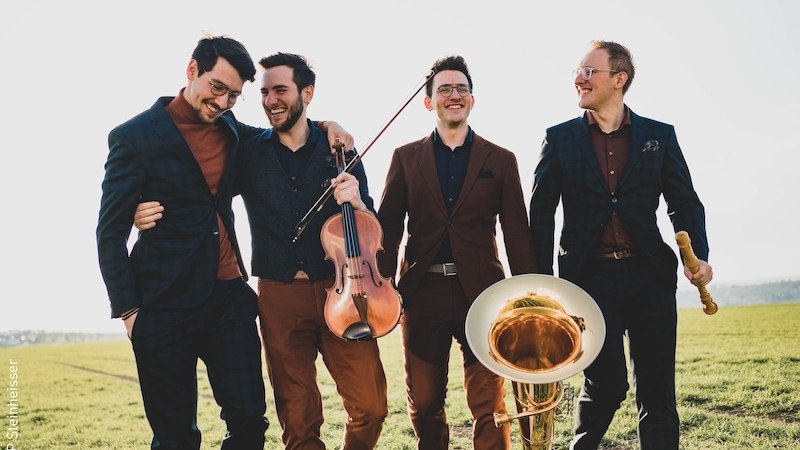 Vier Männer in Anzügen lachen und gehen auf einem Feld. Einer hält eine Geige, ein anderer ein Horn. Sie wirken fröhlich und entspannt., © Stadt Kornwestheim Kulturmanagement Vier Männer in Anzügen lachen und gehen auf einem Feld. Einer hält eine Geige, ein anderer ein Horn. Sie wirken fröhlich und entspannt., © Stadt Kornwestheim Kulturmanagement