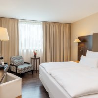 Modernes Doppelzimmer mit großem Bett, Schreibtisch, Sessel und Fenster mit beigen Vorhängen. Ein Fernseher und eine Stehlampe sind ebenfalls vorhanden., © NH Hotel Stuttgart Airport