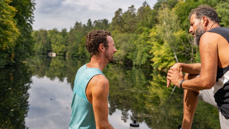 Zwei Männer sitzen am Ufer eines Sees, umgeben von grünen Bäumen, und unterhalten sich. Einer hält eine Wasserflasche., © SMG, Martina Denker Zwei Männer sitzen am Ufer eines Sees, umgeben von grünen Bäumen, und unterhalten sich. Einer hält eine Wasserflasche., © SMG, Martina Denker