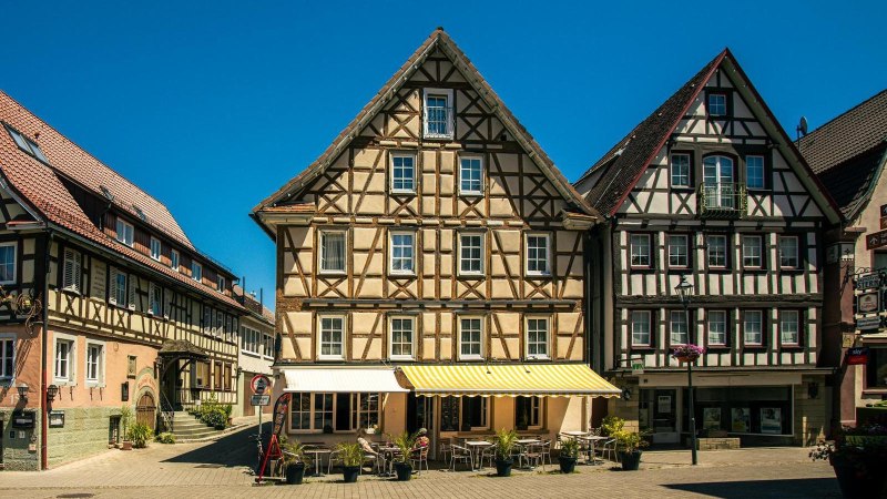 Fachwerkhäuser am Marktplatz von Murrhardt unter klarem, blauem Himmel. Vor den Gebäuden stehen Tische und Stühle eines Cafés., © SMG, Sarah Schmid Fachwerkhäuser am Marktplatz von Murrhardt unter klarem, blauem Himmel. Vor den Gebäuden stehen Tische und Stühle eines Cafés., © SMG, Sarah Schmid