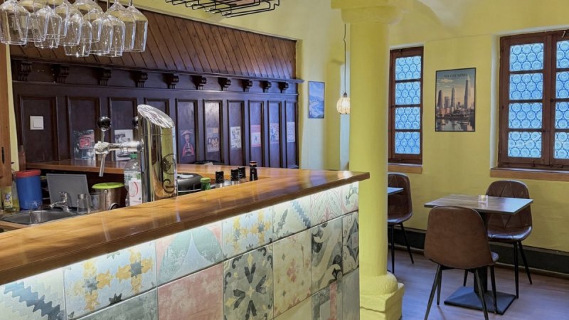 Ein gem&uuml;tliches Restaurant mit gelben W&auml;nden, einer Bar mit bunten Fliesen und h&auml;ngenden Weingl&auml;sern. Im Hintergrund sind Tische und St&uuml;hle zu sehen.