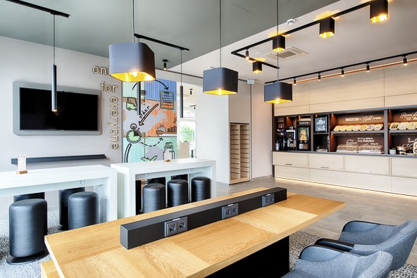 Moderner Frühstücksbereich mit Holztischen, schwarzen Hockern und einer Frühstücksbar im Hintergrund., © B&B Hotel Stuttgart-Neckarhafen