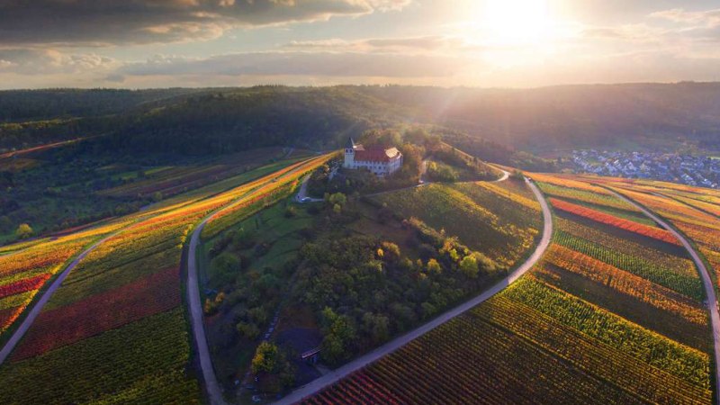 Luftaufnahme des Michaelsbergs, umgeben von bunten Weinbergen im Sonnenuntergang. Eine Kirche steht auf dem Hügel, umgeben von Wald und Feldern., © Neckar-Zaber-Tourismus e.V