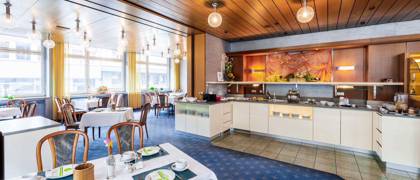 Ein heller Frühstücksraum mit Holzelementen, gedeckten Tischen und einem Buffet. Große Fenster lassen viel Licht herein., © TOMAS