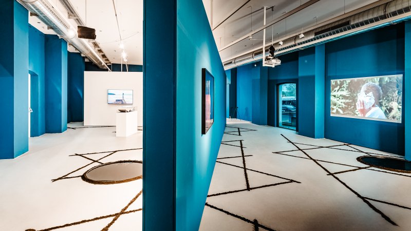 Moderne Galerie mit blauen Wänden, Videoinstallationen und geometrischen Mustern auf dem Boden. Industrielles Design mit sichtbaren Lüftungsrohren., © Ifa Galerie Stuttgart Moderne Galerie mit blauen Wänden, Videoinstallationen und geometrischen Mustern auf dem Boden. Industrielles Design mit sichtbaren Lüftungsrohren., © Ifa Galerie Stuttgart