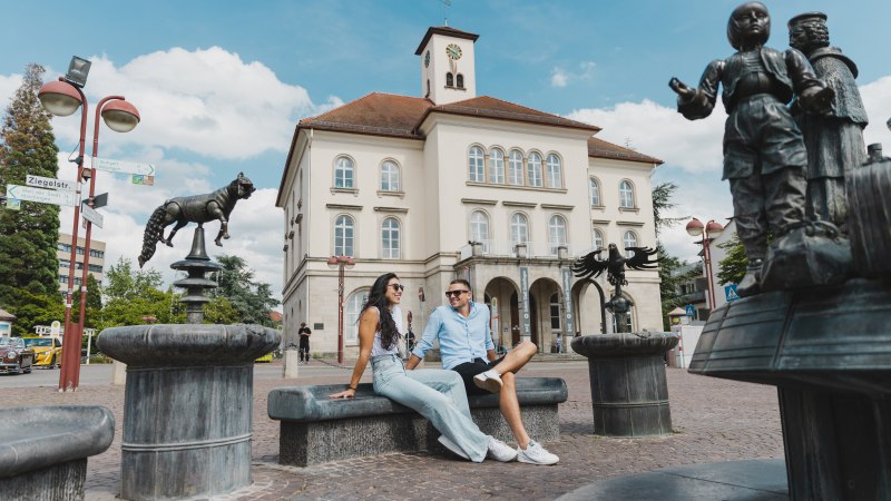 Zwei Personen sitzen vor einem historischen Gebäude in Sindelfingen. Im Vordergrund sind Skulpturen und ein Brunnen zu sehen. Der Himmel ist blau mit Wolken., © benjaminknoblauch.de