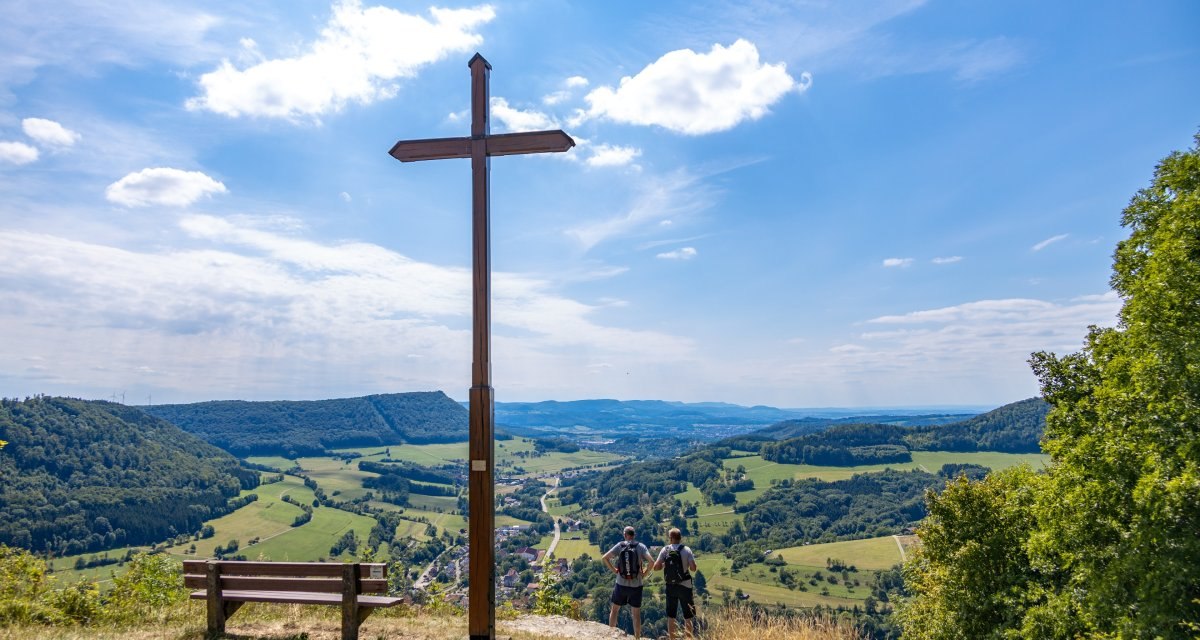 Gipfelkreuz auf einem H&uuml;gel mit Bank, zwei Personen blicken auf die weite Landschaft. Der Himmel ist blau mit einigen Wolken., &copy; Foto Thomas Zehnder