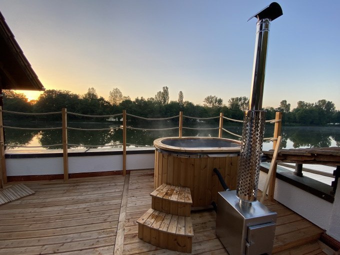 Holzterrasse mit Hot Tub, daneben ein Ofen. Im Hintergrund ein Fluss und Bäume bei Sonnenuntergang., © Cool-Tours StattReisen Holzterrasse mit Hot Tub, daneben ein Ofen. Im Hintergrund ein Fluss und Bäume bei Sonnenuntergang., © Cool-Tours StattReisen