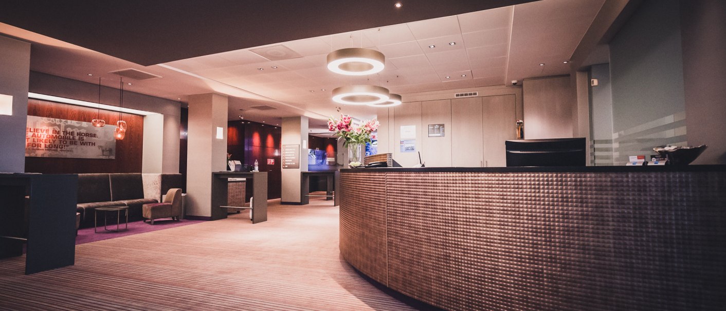 Eine moderne Hotel-Lobby mit einem geschwungenen Empfangstresen, stilvoller Beleuchtung und Sitzbereich. Blumenarrangement auf dem Tresen., © Abacco Hotels GmbH Eine moderne Hotel-Lobby mit einem geschwungenen Empfangstresen, stilvoller Beleuchtung und Sitzbereich. Blumenarrangement auf dem Tresen., © Abacco Hotels GmbH