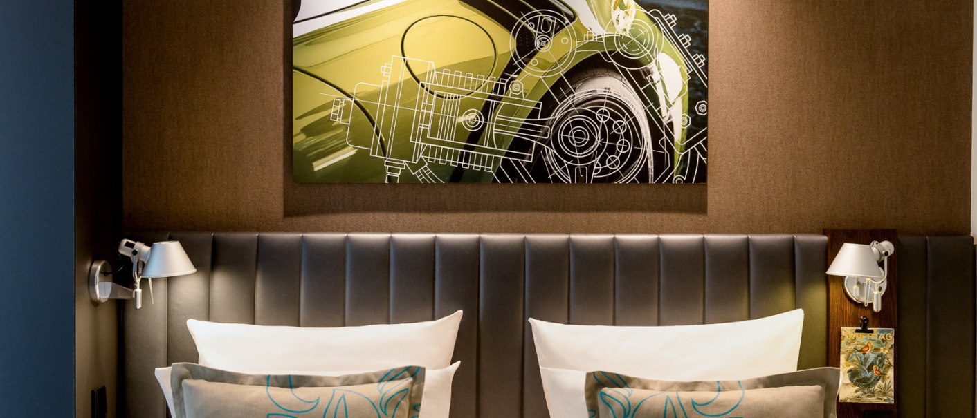 Ein Hotelzimmer mit einem großen Kunstwerk eines Autos über dem Bett. Das Bett hat graue Kissen und eine gepolsterte Rückwand., © TOMAS