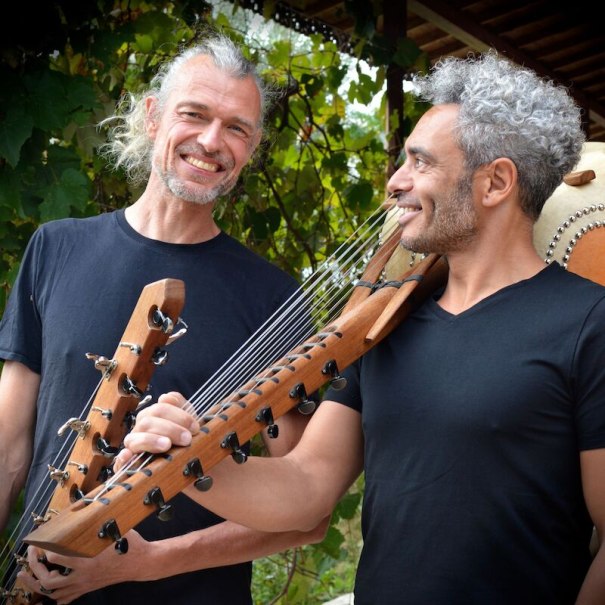 Zwei M&auml;nner mit Saiteninstrumenten l&auml;cheln sich an. Sie stehen vor einer gr&uuml;nen, bl&auml;ttrigen Kulisse und tragen schwarze T-Shirts., &copy; Interessengemeinschaft Kultur Sindelfingen / B&ouml;blingen e.V.