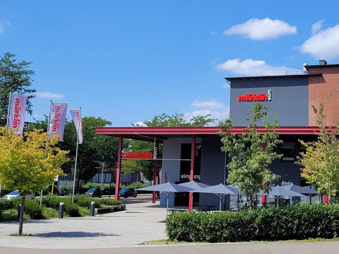 Das M&auml;rklineum-Geb&auml;ude mit rotem Schriftzug und Eingang, umgeben von B&auml;umen und Flaggen. Der Himmel ist blau mit wenigen Wolken., &copy; M&auml;rklin Spielwaren Vertriebs-GmbH