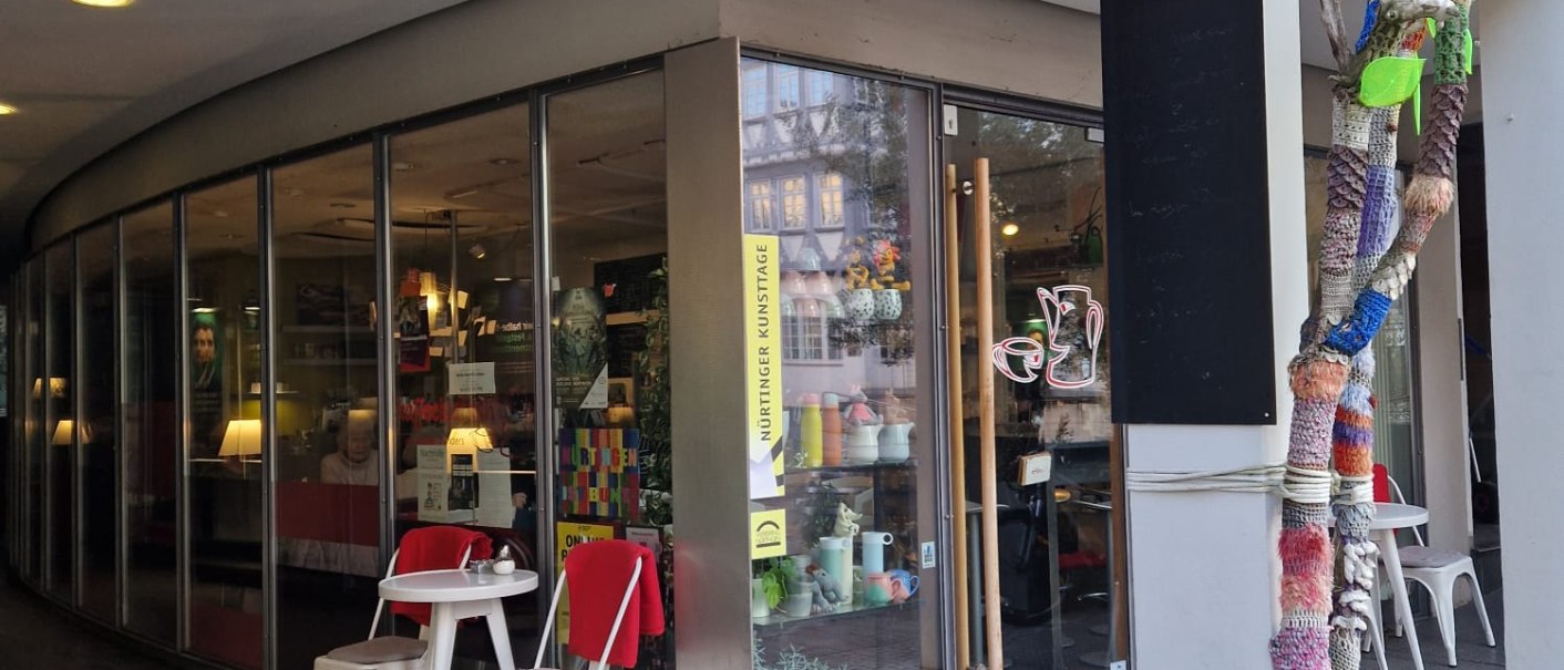 Außenansicht eines Cafés mit Glastüren und einem kleinen Tisch mit zwei Stühlen. Ein Baumstamm ist bunt umstrickt., © cStadtNuertingen Außenansicht eines Cafés mit Glastüren und einem kleinen Tisch mit zwei Stühlen. Ein Baumstamm ist bunt umstrickt., © cStadtNuertingen