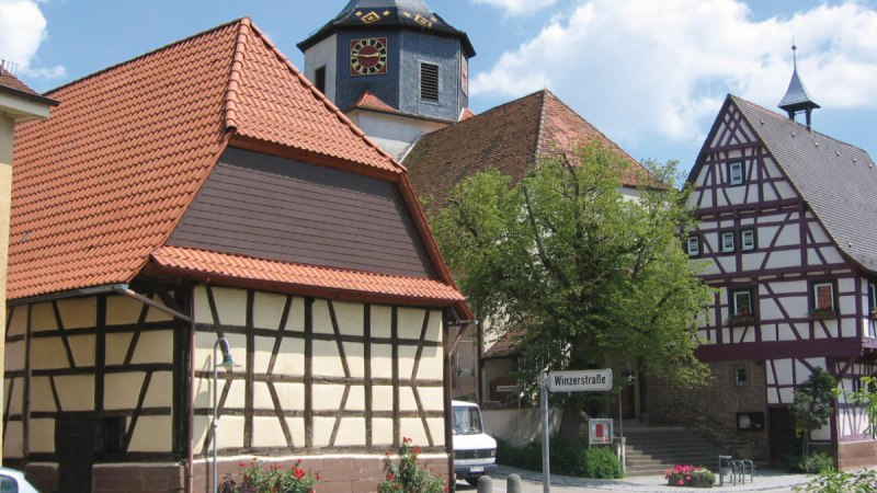 Fachwerkhäuser in Gündelbachs Ortsmitte, mit einem Kirchturm im Hintergrund. Ein Straßenschild zeigt 'Winzerstraße'. Sonniger Tag mit blauem Himmel., © Land der 1000 Hügel - Kraichgau-Stromberg