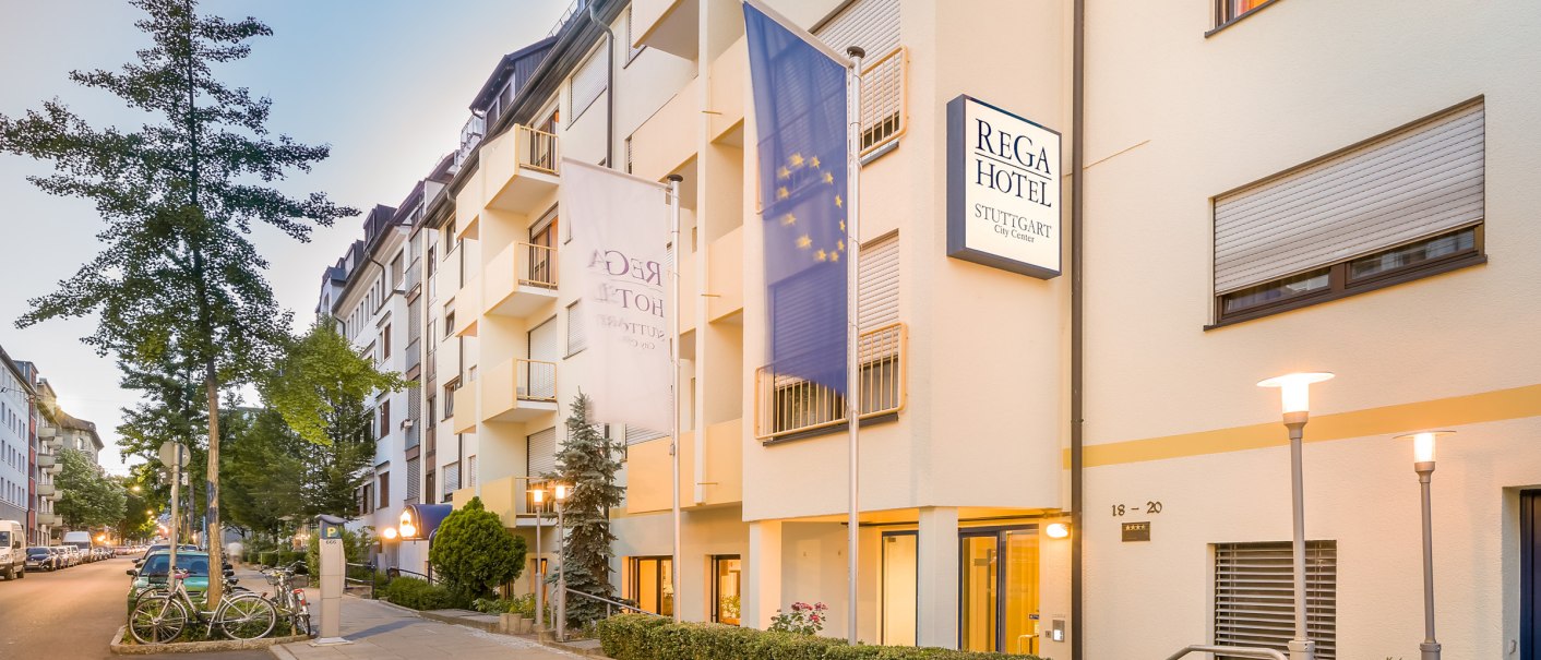 Außenansicht des Rega Hotels in Stuttgart. Mehrstöckiges Gebäude mit Fahnen und beleuchteten Straßenlaternen. Ruhige Straße mit geparkten Autos., © TOMAS