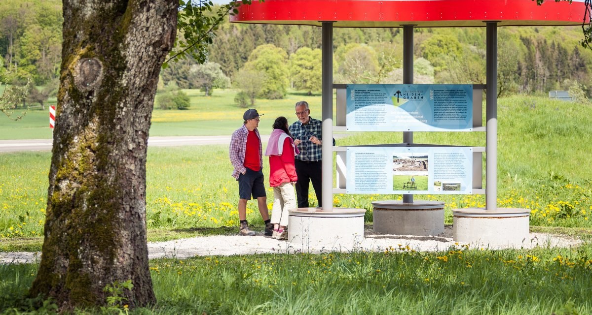 Drei Personen stehen vor einem Infopavillon auf einer gr&uuml;nen Wiese. Der Pavillon hat ein rotes Dach und Informationstafeln. Im Hintergrund sind B&auml;ume zu sehen., &copy; hochgehberge
