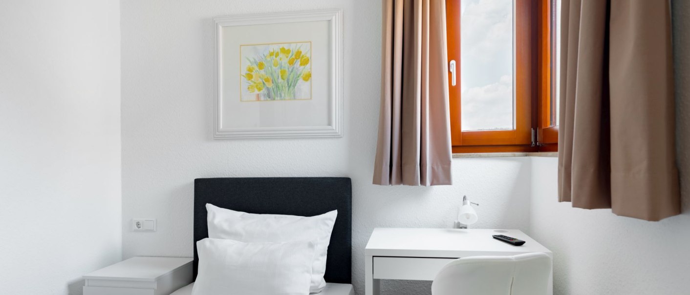 Helles Einzelzimmer mit einem Bett, einem weißen Schreibtisch und einem Fenster mit braunen Vorhängen. An der Wand hängt ein Bild mit gelben Blumen., © Hotel Olgaeck
