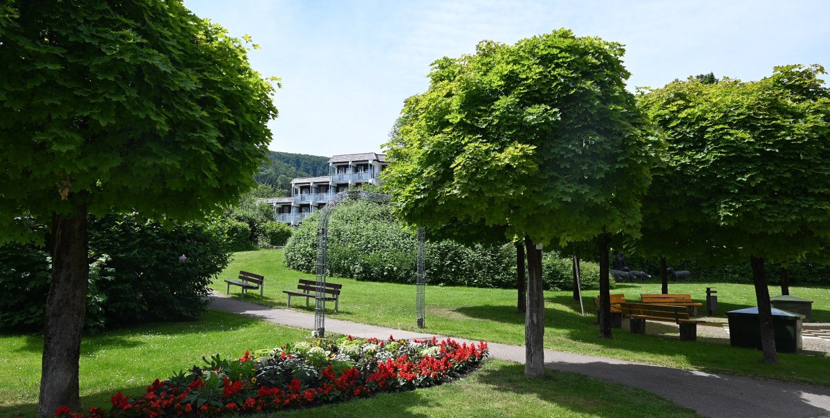 Ein grüner Park mit Blumenbeeten, Bänken und Bäumen. Im Hintergrund ist ein mehrstöckiges Gebäude zu sehen.