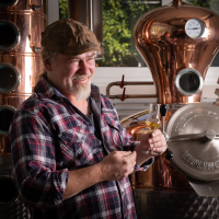Mann in kariertem Hemd hält ein Glas mit goldenem Getränk vor einer Kupferdestille. Er lächelt und trägt eine Mütze. Im Hintergrund sind Fenster und Bäume sichtbar., © Brau- & Brennwerkstatt