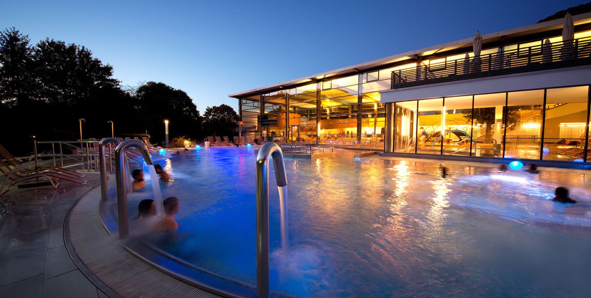 Die AlbThermen bei Nacht, beleuchtet mit einem Außenpool. Menschen entspannen im Wasser, während das Gebäude warm erleuchtet ist., © AlbTherme Bad Urach