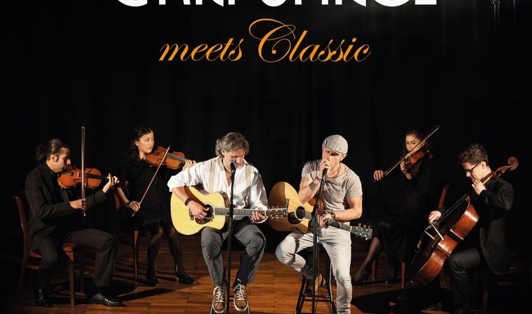 Zwei Musiker mit Gitarren sitzen auf einer B&uuml;hne, begleitet von einem Streichquartett. Oben steht 'Simon & Garfunkel Tribute Duo meets Classic'., &copy; Graceland
