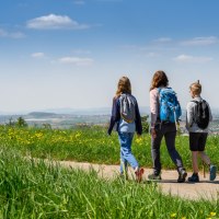 Drei Personen wandern auf einem Weg durch eine grüne Wiese mit gelben Blumen, im Hintergrund eine weite Landschaft unter blauem Himmel., © Stadtverwaltung Winnenden Drei Personen wandern auf einem Weg durch eine grüne Wiese mit gelben Blumen, im Hintergrund eine weite Landschaft unter blauem Himmel., © Stadtverwaltung Winnenden