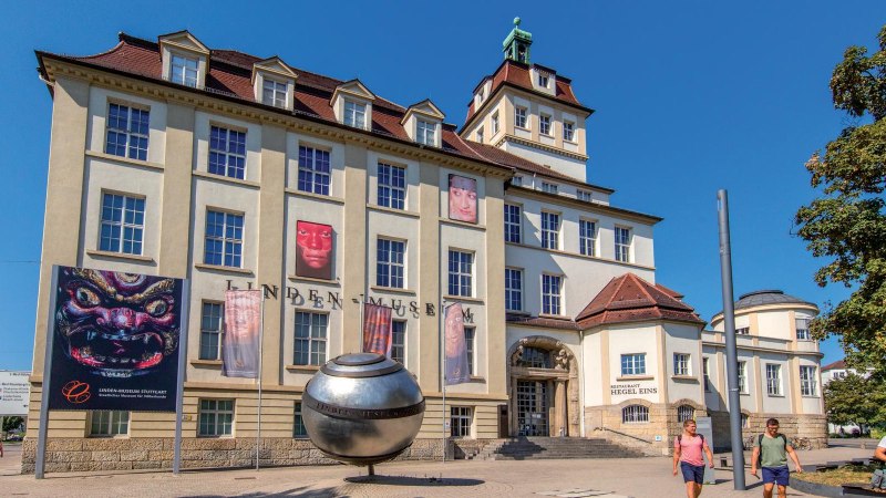 Das Linden-Museum in Stuttgart bei sonnigem Wetter. Vor dem Gebäude sind Plakate und eine große silberne Kugel zu sehen. Menschen spazieren vorbei., © Stuttgart-Marketing GmbH, Achim Mende